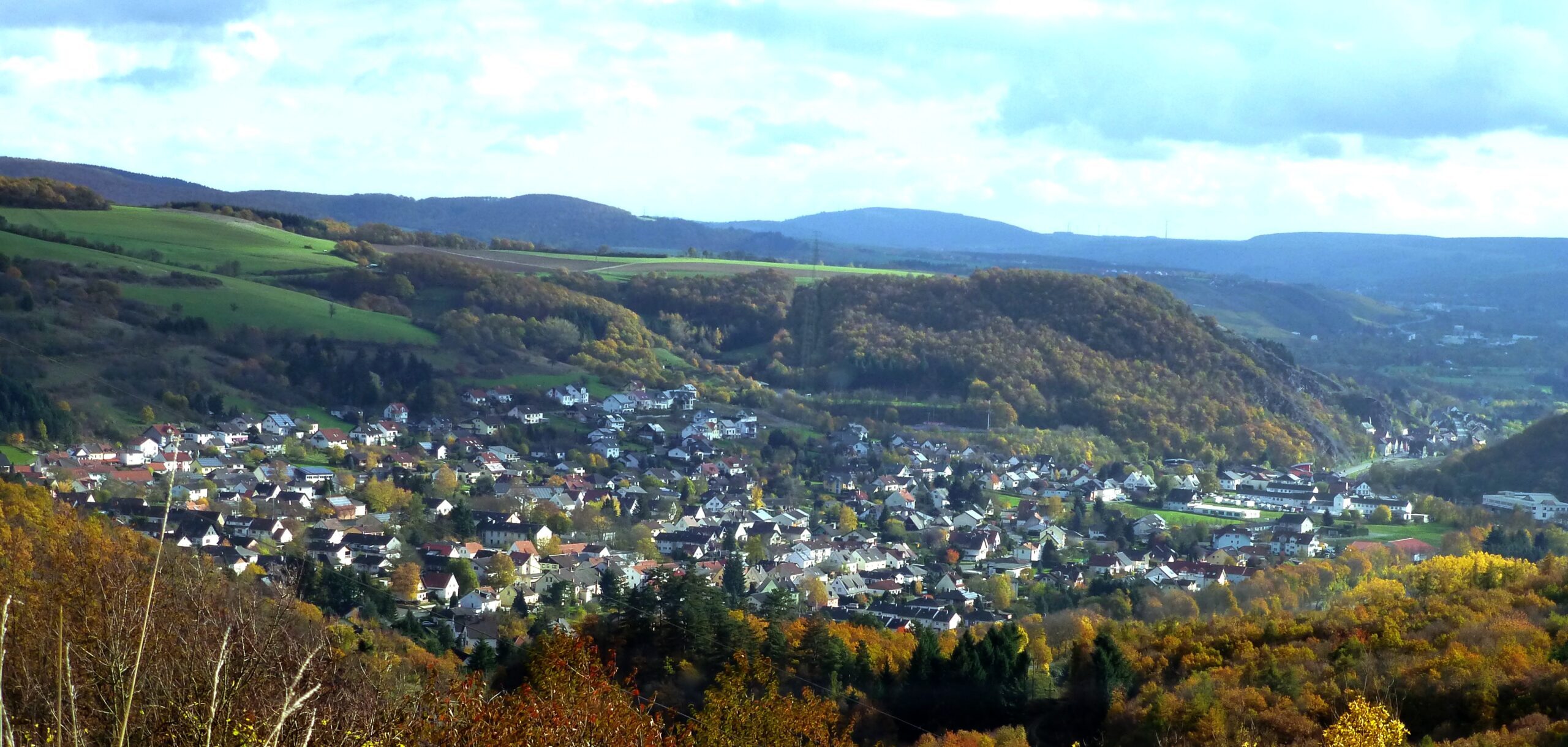 Panoramablick auf Simmertal mit Wohngebiet, bewaldeten Hügeln, Feldern und herbstlicher Landschaft unter bewölktem Himmel. Foto: giggel, Wikimedia Commons, lizenziert unter CC BY 3.0