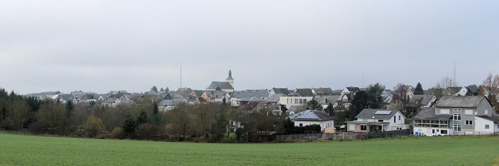 Panoramablick auf die Ortsgemeinde Mörsdorf mit Feldern im Vordergrund und einer geschlossenen Bebauung im Hintergrund unter bedecktem Himmel. Foto-Credit Urheber: Colling-architektur Lizenz: Creative Commons Attribution-Share Alike 3.0 Unported (CC BY-SA 3.0)