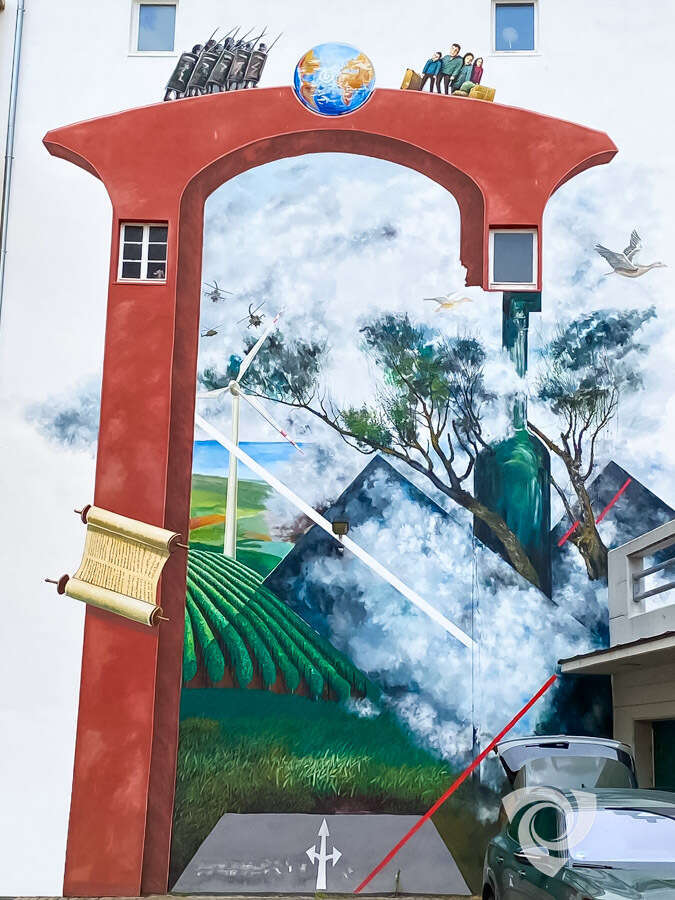Die Aufnahme zeigt einen zentralen Teil des Wandgemäldes „Ein Tor zur Welt“ in Alzey. Zu sehen sind Windenergieanlagen in einer Landschaft mit Weinbergen sowie ein großer roter Torbogen, der das Motiv prägt. Oberhalb des Torbogens befinden sich eine Erdkugel und weitere symbolische Darstellungen. Im rechten Bildbereich sind eine große Weinflasche und Bäume abgebildet. Das Kunstwerk verbindet regionale Elemente Rheinhessens mit Themen wie Umwelt, Nachhaltigkeit und globaler Vernetzung.